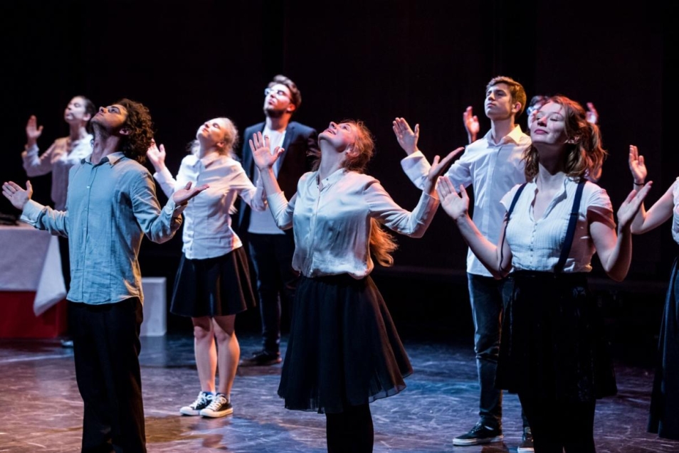 Grupa młodzieży biorąca udział w spektaklu teatralnym. Młodzi artyści ustawieni są w trzech rzędach, mają odchylone głowy i uniesione ręce.