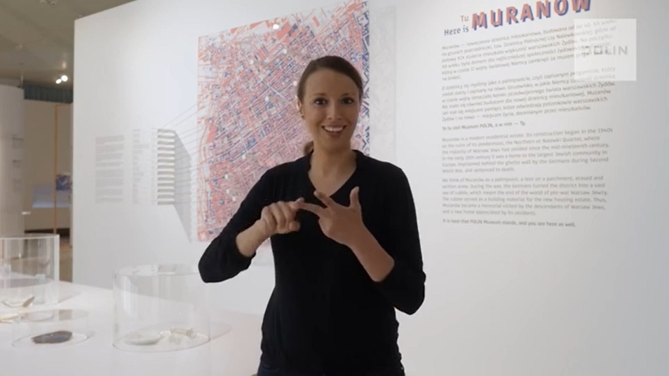 Tłumaczka Polskiego Języka Migowego stoi w przestrzeni wystawy Tu Muranów. Za plecami ma planszę ze wstępem do wystawy i planem Muranowa.