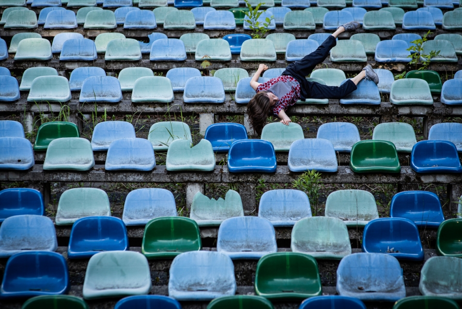 Rzędy niebieskich stadionowych krzesełek, na jednym z nich, z głową w dół, leży jedna osoba