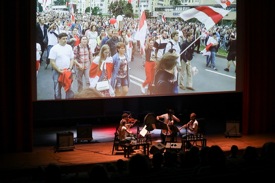 Scena - kwartet smyczkowy - nad sceną ekran, na którym wyświetlane jest wideo z ludźmi, którzy protestują przeciw prześladowaniom Białorusinów na ulicy w Mińsku