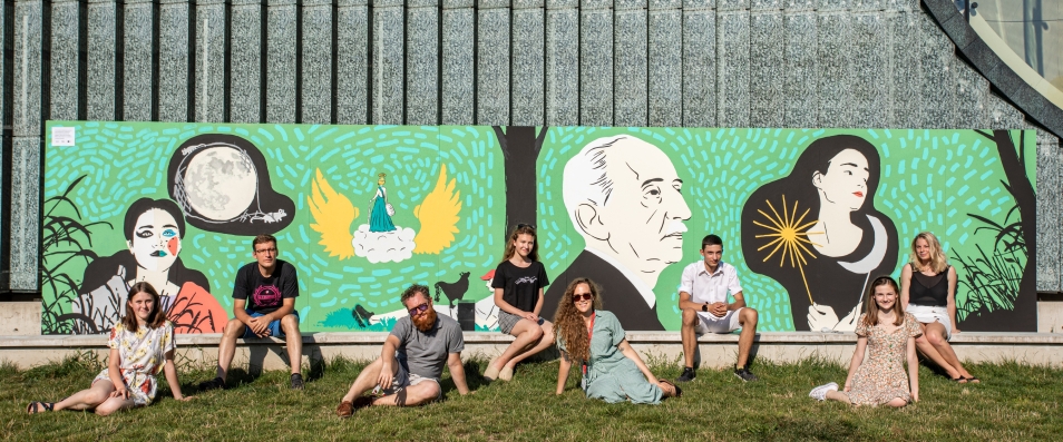 Mural na Łące Leśmiana przedstawia głowę Bolesława Leśmiana i osoby związane z jego twórczością. Przed muralem na trawie i murku siedza osoby, które tworzyły mural