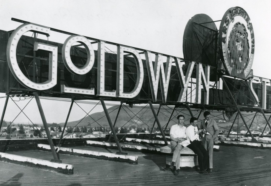 Czarno białe zdjęcie przedstawia ogromny neon "Goldwyn". Pod jego konstrukcją siedzi trzech mężczyzn.
