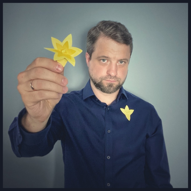 In Polish: męzczyzna w czarnej koszuli ma przypięty do piersi papierowy żonkil, symbol akcji Żonkile. Drugi trzyma w wyciągniętej w stronę obiektywu ręce.