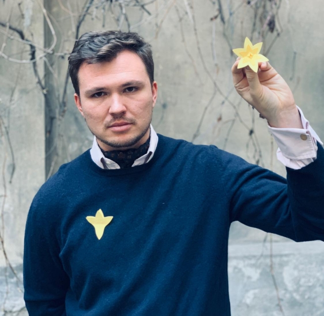 In Polish: Stefan Thomas w granatowym swetrze. Ma przypięty do swetra papierowy żonkil, w podniesionej w górę dłoni trzyma drugi żonkil.