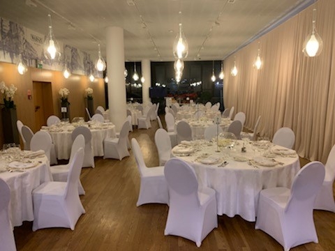 Wnętrze restauracyjne odświętnie zastawione - wokół okrągłych stołów na 8 osób, które są nakryte białymi obrusami, stoja krzesła w eleganckich białych pokrowcach. Wnętrze jest nastrojowo oświetlone.