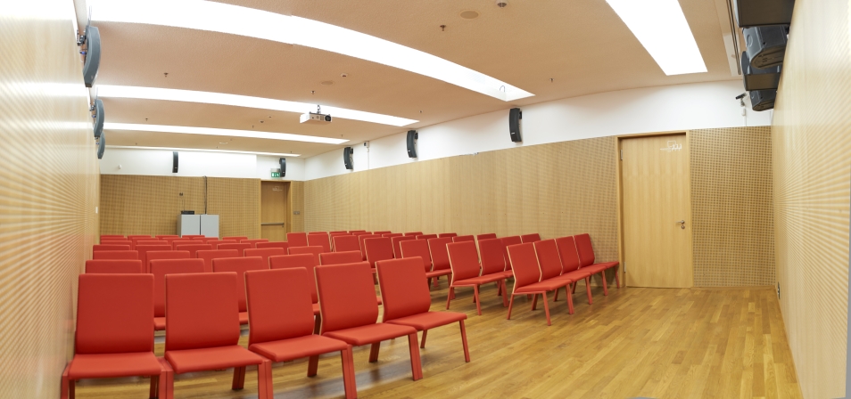 Sala konferencyjna, w której rzędami stoją czerwone krzesła. Sala jest pusta.