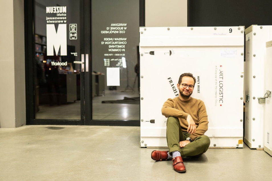 Piotr Skłodkowski siedzi na podłodze oparty o skrzynię z zabytkami, na kolenie jednej z nóg opiera rękę.