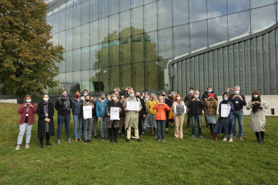 Grupa osób w jesiennych ubraniach stoi przed budynkiem Muzeum POLIN. Część trzyma transparenty, część dłonie złożone w symbol serca. Apel ws. kryzysu uchodźczego na granicy Polski i Białorusi