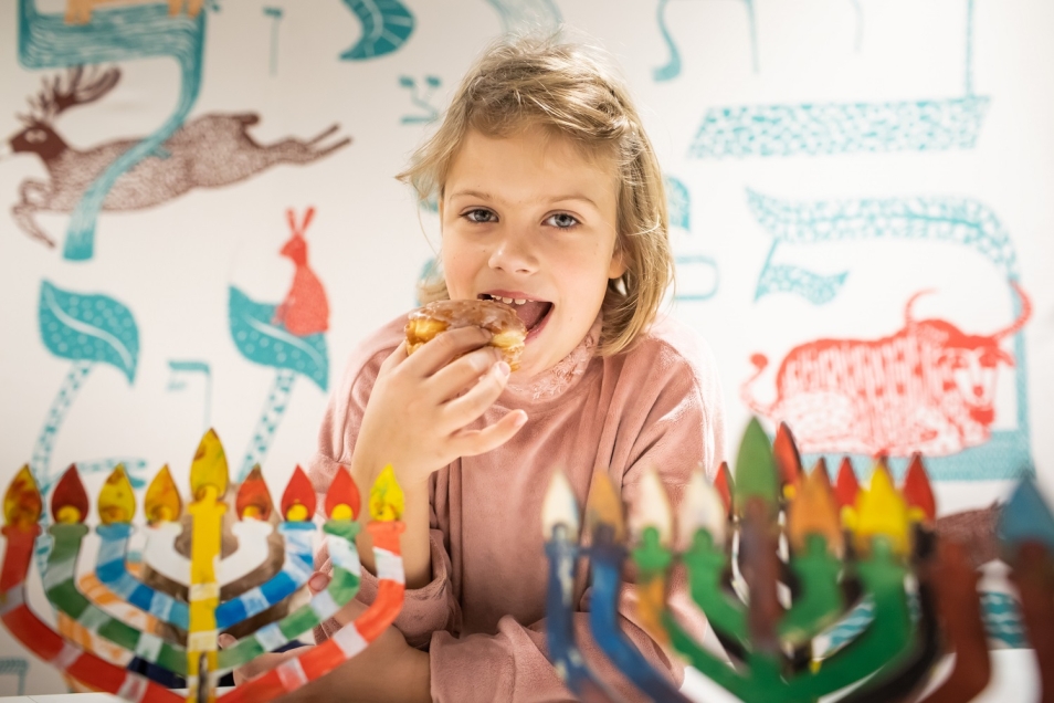 Dziewczynka je pączka. Przed nią dwa modele kolorowych świeczników.