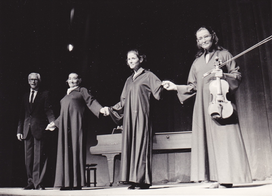 Czarno-biała fotografia. Lin Jaldati Ensemble 1984. Jeden starszy mężczyzna w garniturze i trzy kobiety w długich szatach stoją na scenie, trzymając się za ręce. Kobieta, która stoi z prawej strony, trzyma skrzypce.
