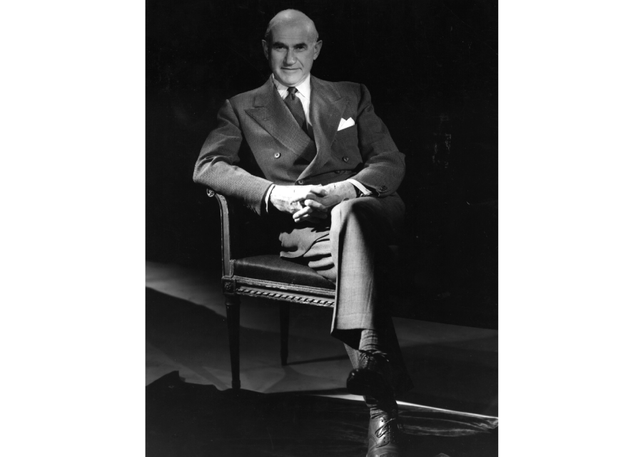 amuel Goldwyn. Czarno-białe zdjęcie portretowe. Starszy, łysy pan w garniturze siedzi na krześle. Ma splecione dłonie.