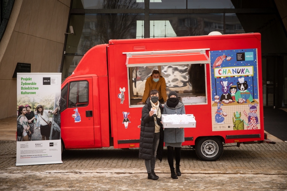 Chanukobus przed Muzeum POLIN. Czerwony autobus z okienkiem. Przed nim stoją dwie kobiety z pączkami.