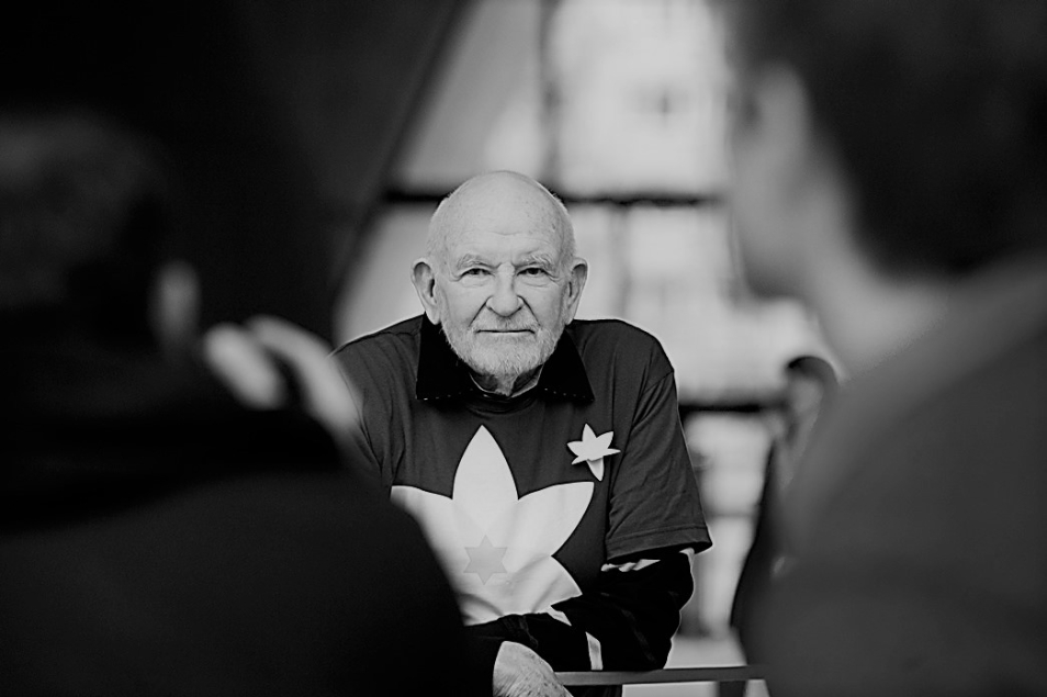 Portret Janusza Czamarskiego - widać go zza ramion osób z pierwszym planie. Ma na sobie ciemna koszulkę z żółtym żonkilem.