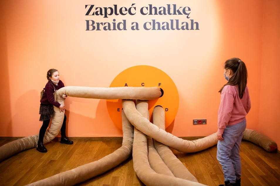 Dwie dziewczynki próbują zapleść chałkę na wystawie "Od kuchni. Żydowska kultura kulinarna".