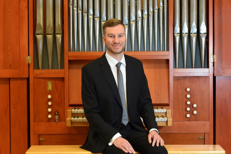 Jakub Stefek - uśmiechnięty mężczyzna w krawacie i garniturze pozuje na tle organów.