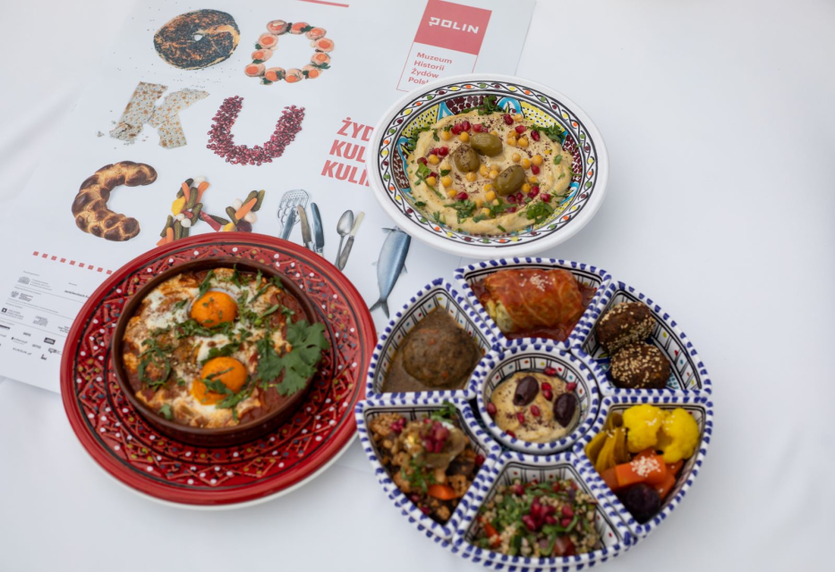 Trzy miski z potrawami kuchni żydowskiej - hummusem, mezze i szakszuką. Obok ulotka wystawy "Od kuchni. Żydowska kultura kulinarna".