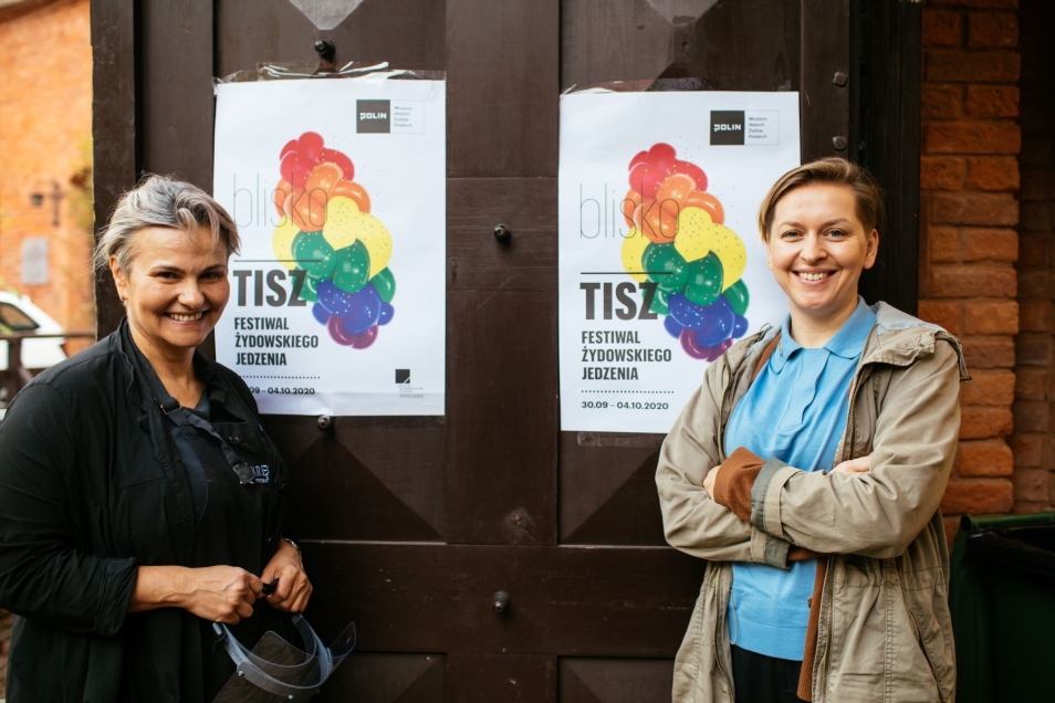 Dwie kobiety stoją przy drzwiach z plakatami Tisz festiwalu 2020. Obie uśmiechają się.