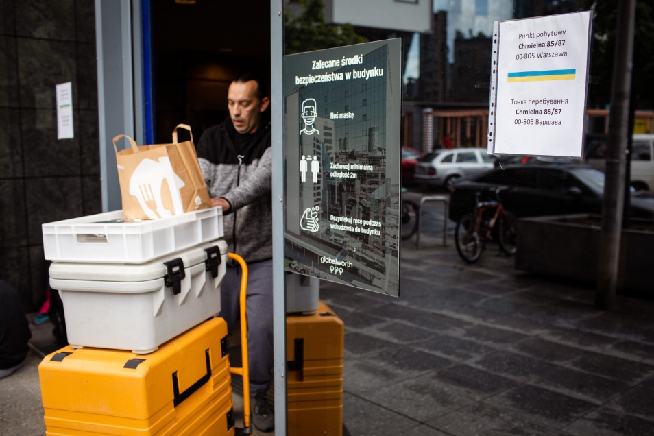 Szklana ściana z drzwiami. Drzwi otwarte. Przechodzi przez nie mężczyzna. Ciągnie wózek transportowy, na którym stoją kontenery z posiłkami dla uchodźców z Ukrainy. ciągnąc wózek