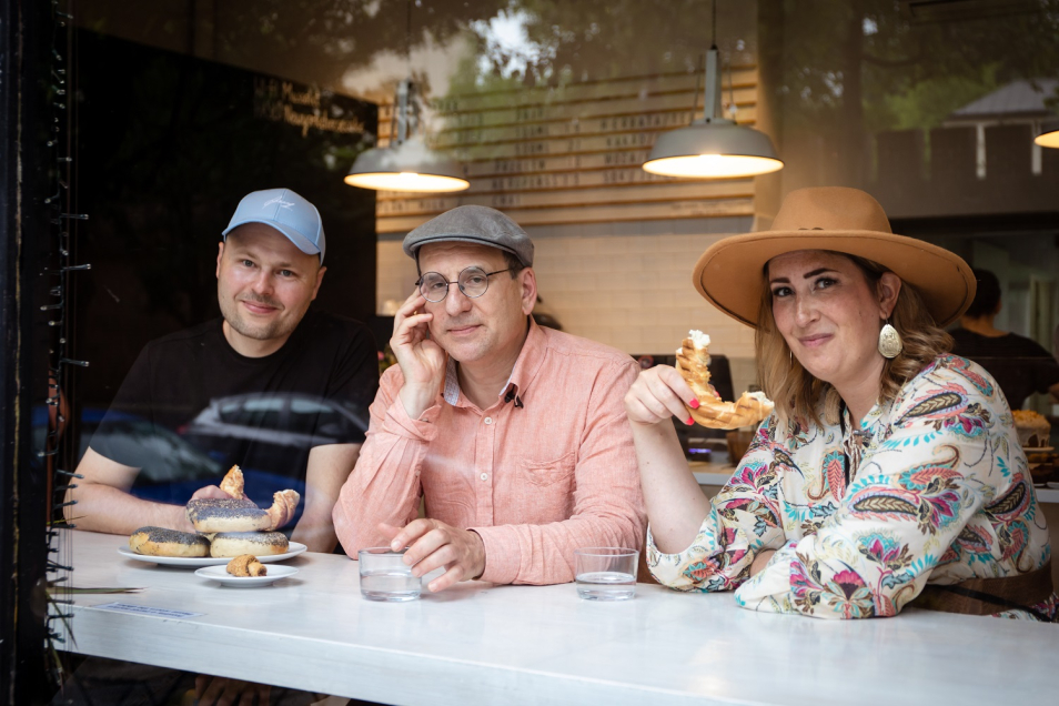 Przy kontuarze w restauracji, przy samej szybie, siedzą trzy osoby i uśmiechają się. Jedzą bajgle.
