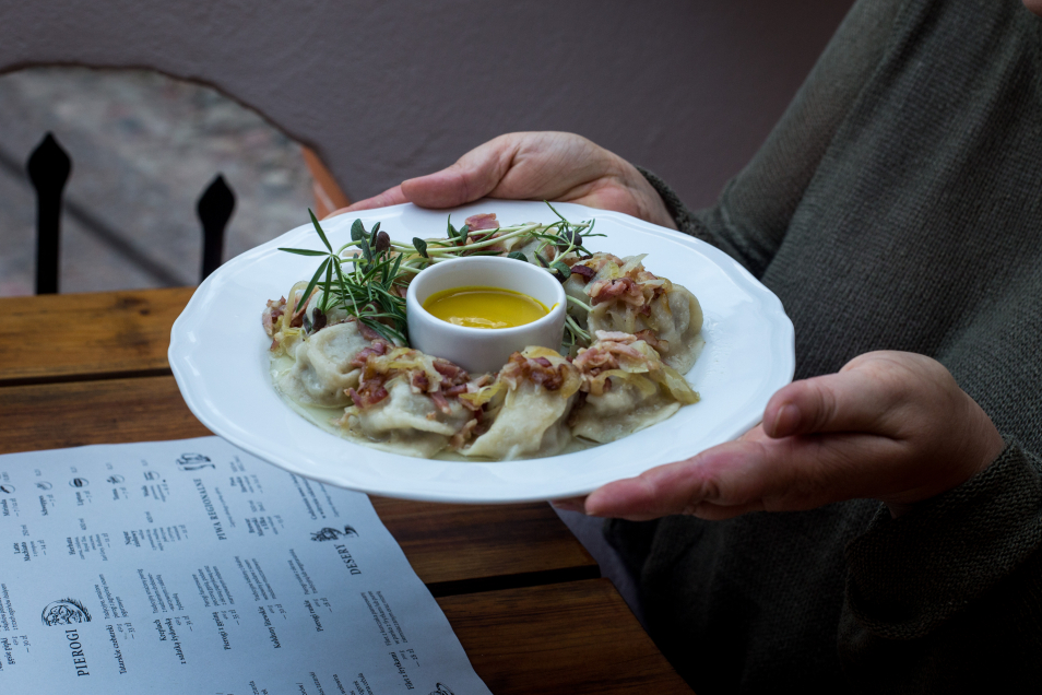 W restauracji - nad blatem stołu ktoś trzyma talerz z pierogami. Między pierogami na środku talerza małe naczynie z sosem.