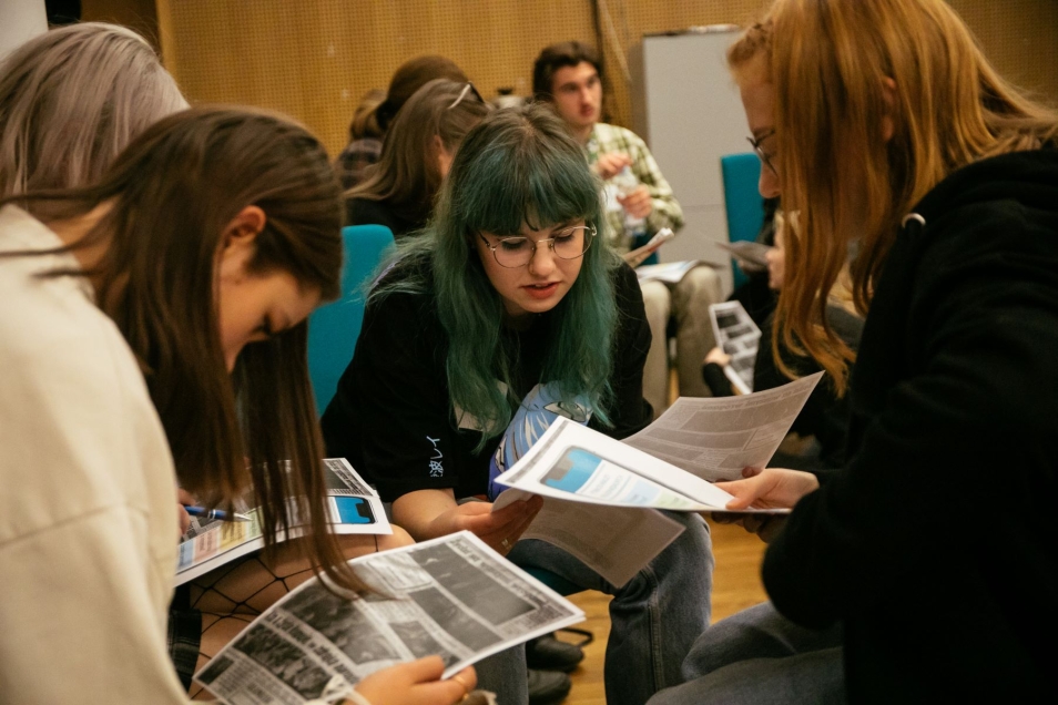 Cztery dziewczyny siedzą razem i czytają coś z kartek, które trzymają przed sobą.