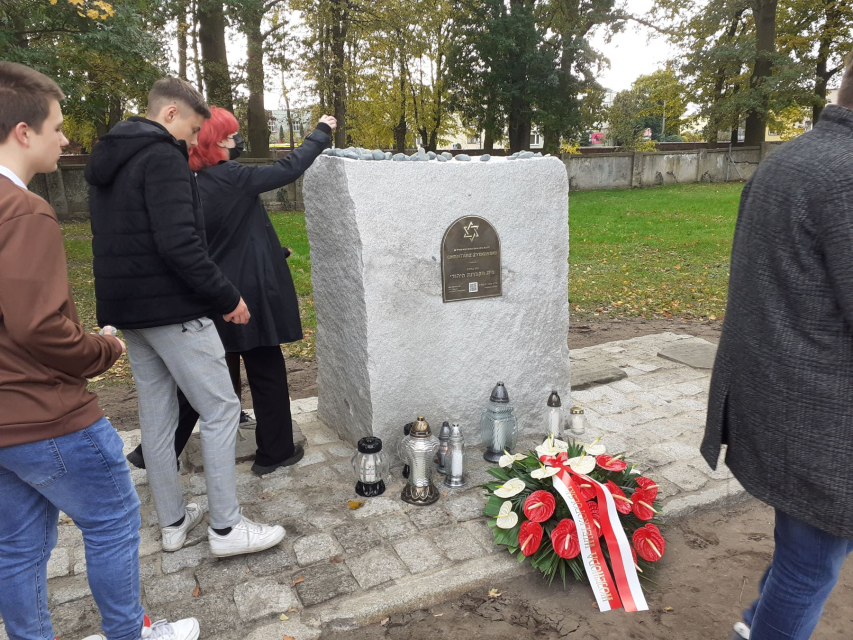Trzy osoby stoją wokół tablicy upamiętniającej cmentarz żydowski w Szamotułach. Kobieta kładzie kamyk na górze wmurowanej w kamień tablicy.