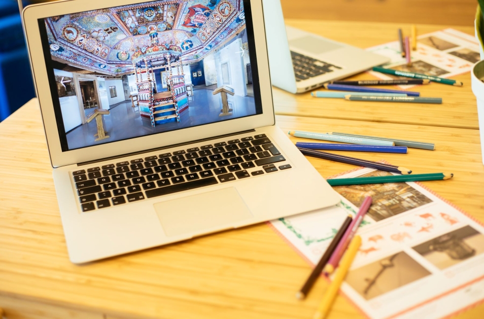 Na stole rozrzucone kredki i zdjęcia. Obok nich stoi laptop. Na ekranie komputera wyświetla się zdjęcie synagogi z wystawy stałej w Muzeum POLIN.