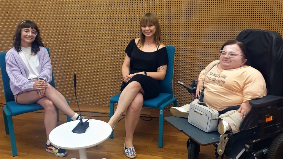 Dwie kobiety siedzą na krzesłach, jedna na wózku inwalidzkim. Przed nimi stolik z mikrofonem.