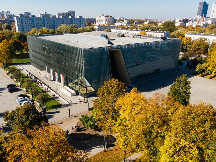Budynek Muzeum POLIN prezentowany z zewnątrz. Wokół niego park, parking, plac i pobliskie bloki.