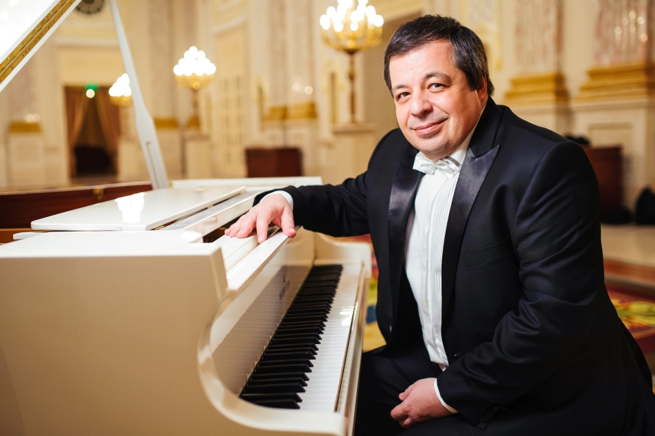 Aleksey Botvinov siedzi przy białym fortepianie w sali ze świecznikami.