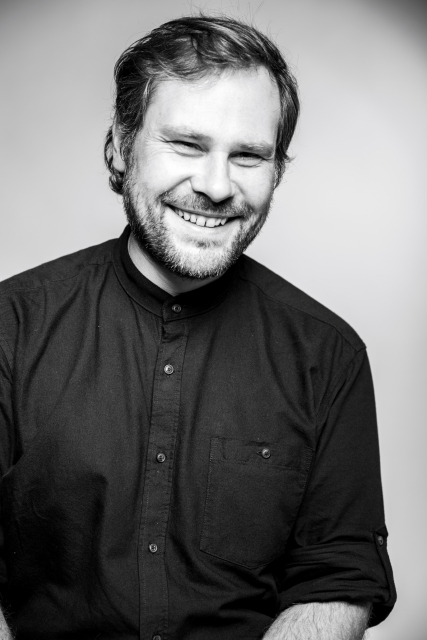 Jakub Jakowicz uśmiecha się. Ma zarost na twarzy i ciemną koszulę.
