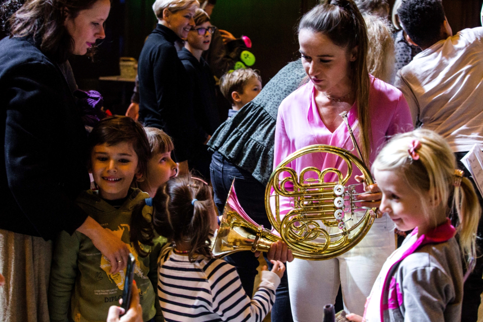 Grupa dzieci stoi wokół kobiety z instrumentem dętym. 