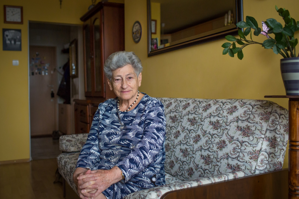 Krystyna Budnicka siedzi na kanapie w mieszkaniu. Ma założone obie ręce na kolanach. Kobieta nosi wzorzystą, biało-granatową bluzkę.