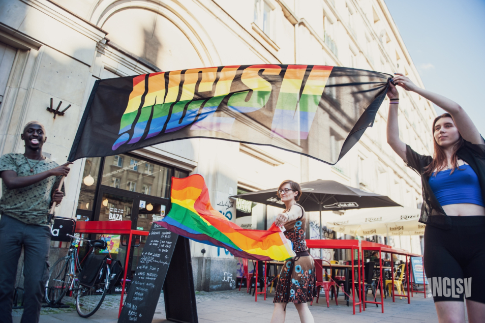 Kobieta i mężczyzna trzymają flagę z napisem "Inclsv". Z tyłu inna kobieta trzyma tęczową flagę.