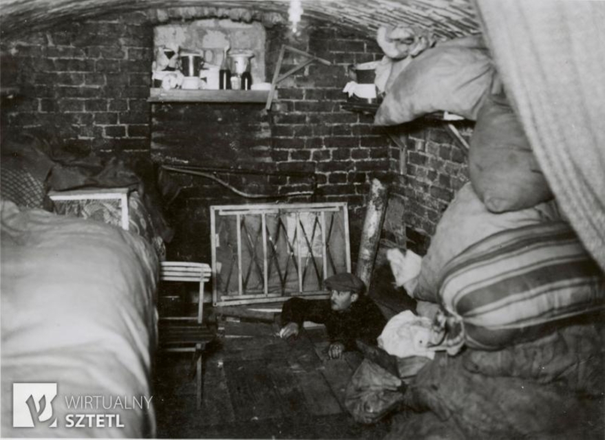Схованка у Варшавському гетто під час повстання 1943 року. Всередині ліжко, мішки, посуд. Всередину через люк входить чоловік у кашкеті.