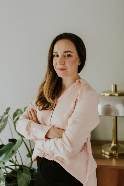Marta Kielak stoi nieco bokiem do nas, ma na sobie jasną bluzkę i ciemne spodnie. Ciemne długie włosy ma przełożone przez prawe ramię