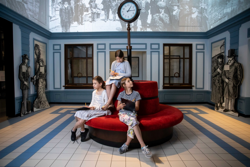 Trzy dziewczynki siedzą na kanapie w galerii "Wyzwania nowoczesności" na wystawie stałej w Muzeum POLIN. W dłoniach trzymają podkładki na kartki i ołówki.