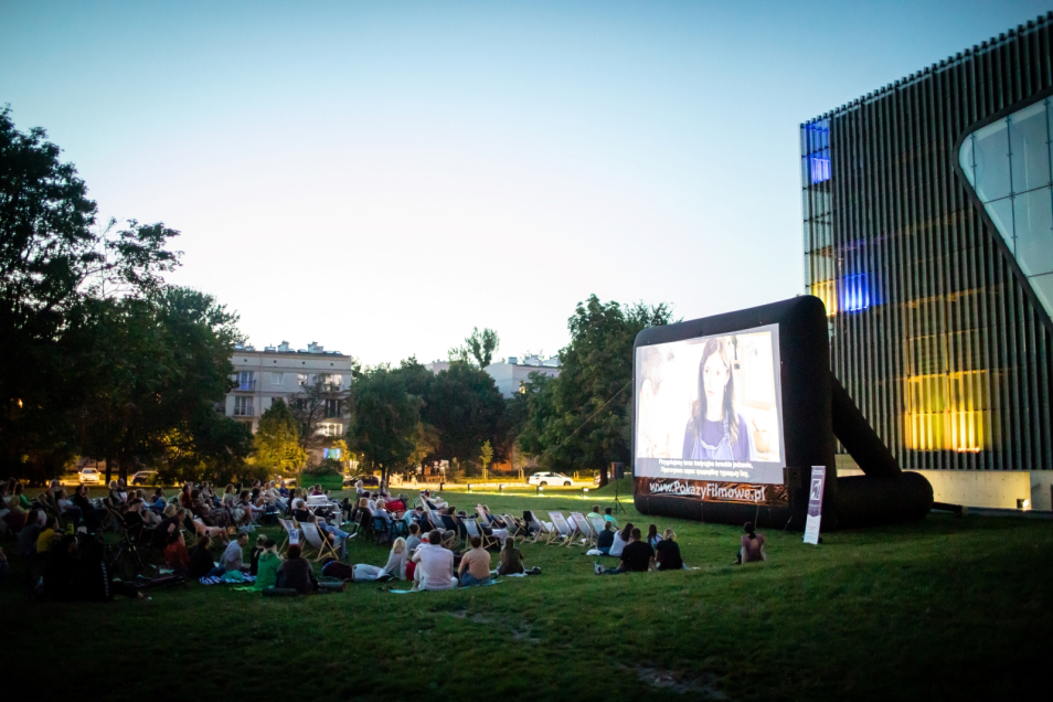 Widzowie siedzą na łące i oglądają film prezentowany na ekranie rozstawionym przy budynku Muzeum POLIN.