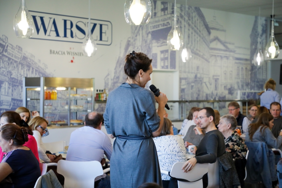 Przewodniczka mówi coś przez mikrofon. Wokół niej przy stolikach siedzą goście restauracji "Warsze".