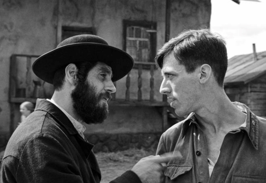 Czarno-biały kadr z filmu "Shttl" - mężczyzna z ciemną brodą i w kapeluszu celuje palcem w pierś drugiego mężczyzny.
