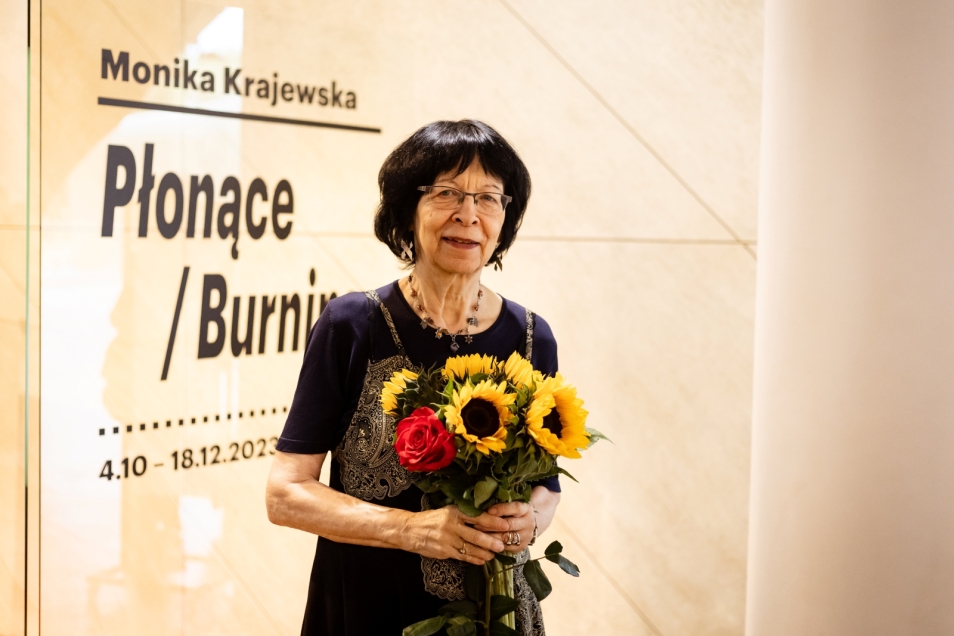 Monika Krajewska trzyma w dłoniach bukiet słoneczników. Za nią na przezroczystej ścianie tytuł jej wystawy "Płonące" / "Burning", 4.10-18.12.2023.