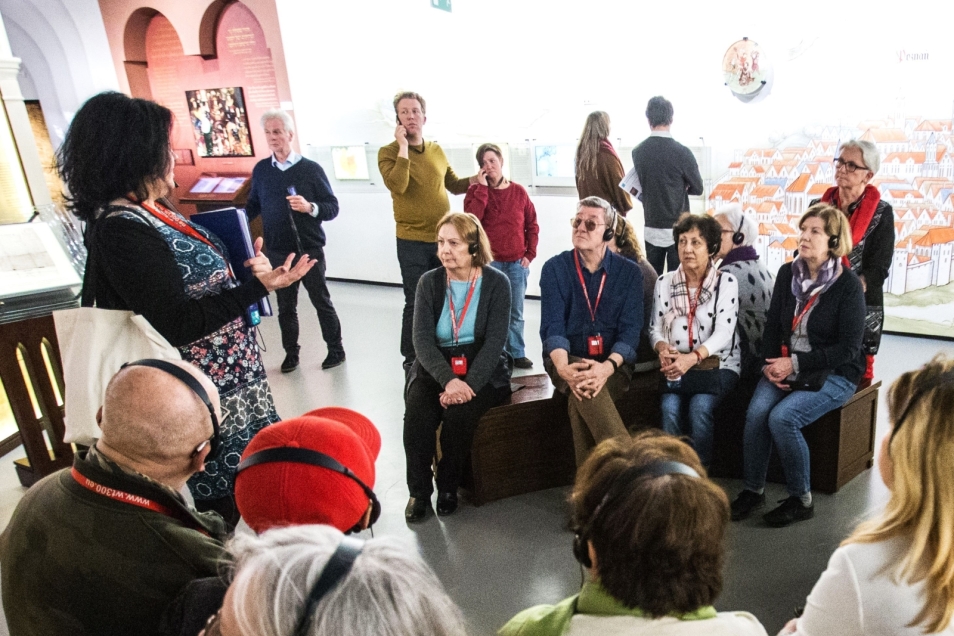 Grupa seniorów siedzi na ławkach na wystawie stałej podczas spaceru z przewodniczką.