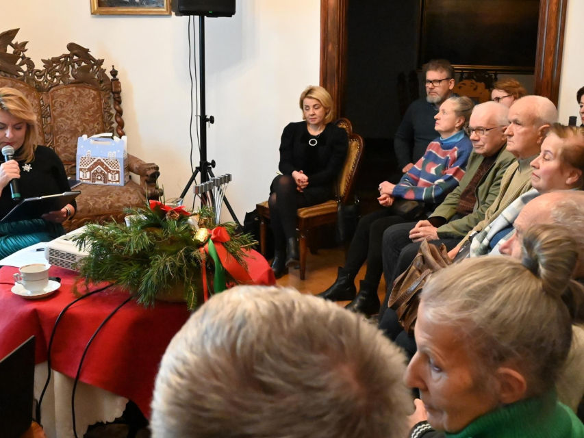 Grupa osób siedzi wokół kobiety mówiącej coś przez mikrofon podczas spotkania chanukowego.