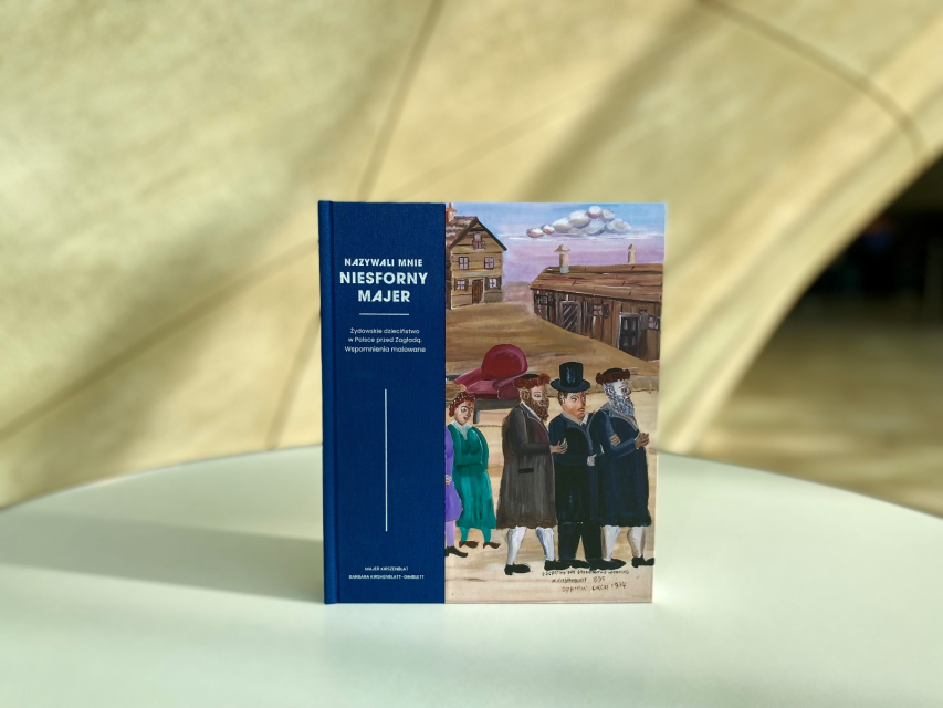 Okładka książki o Majerze Kirszenblacie stoi na tle krzywoliniowej ściany w Muzeum POLIN.