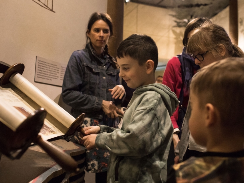 Troje dzieci i kobieta stoją przy zwoju Tory prezentowanym na wystawie stałej Muzeum POLIN.