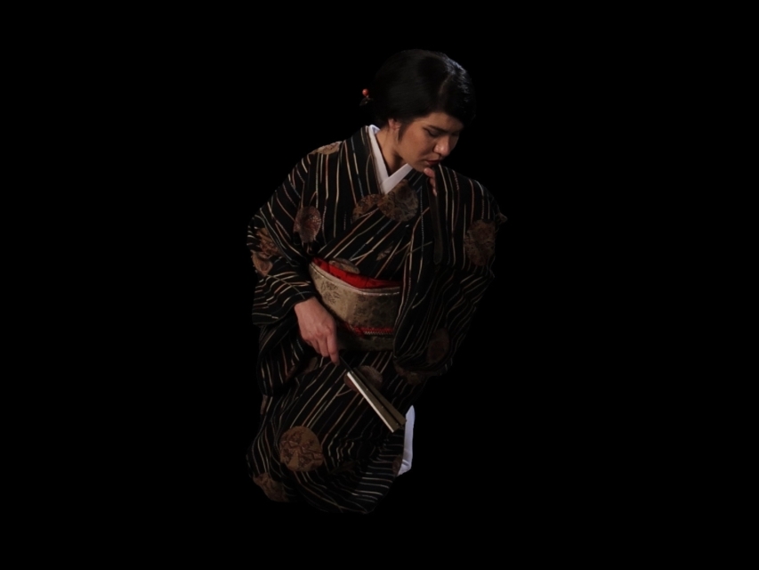 Ktoś, prawdopodobnie kobieta, stoi ubrany w kimono. W dłoni trzyma złożony wachlarz.