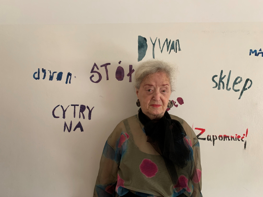 Maya Gordon stoi na tle ściany w Miejskim Ośrodku Sztuki w Gorzowie Wielkopolskim. Na ścianie napisy: cytryna, dzwon, stół, dywan, sklep, zapomnieć.