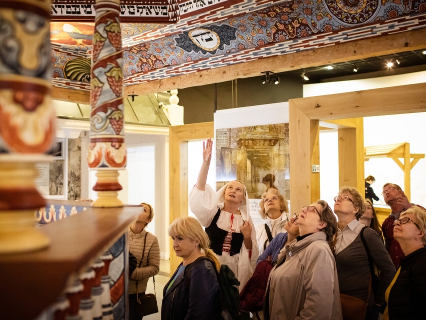 Przewodniczka wskazuje dłonią sklepienie synagogi w Gwoźdźcu znajdującej się na wystawie stałej w muzeum. Wokół niej zgromadzona grupa zwiedzających.