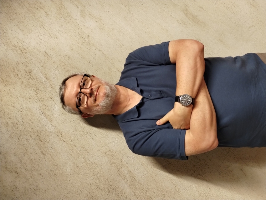 Piotr Kowalik - mężczyzna z brodą i w okularach stoi na tle ściany w Muzeum POLIN.