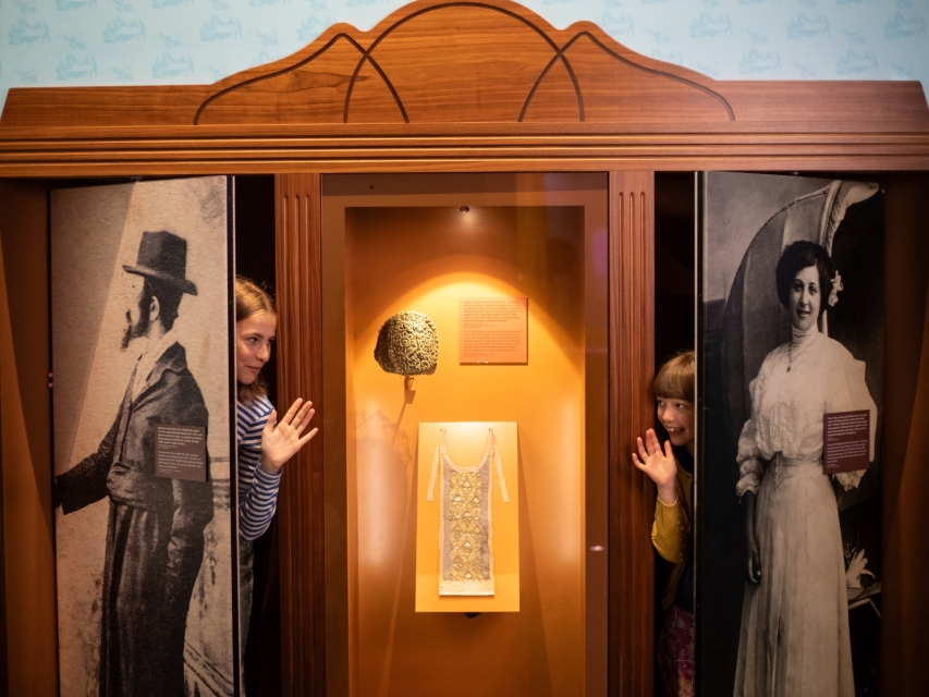 Dwie dziewczyny wychylają się z szafy na wystawie stałej w Muzeum POLIN. Na jednych drzwiach przedstawiona jest postać Żyda w kapeluszu, na drugich - kobieta w białej sukni.